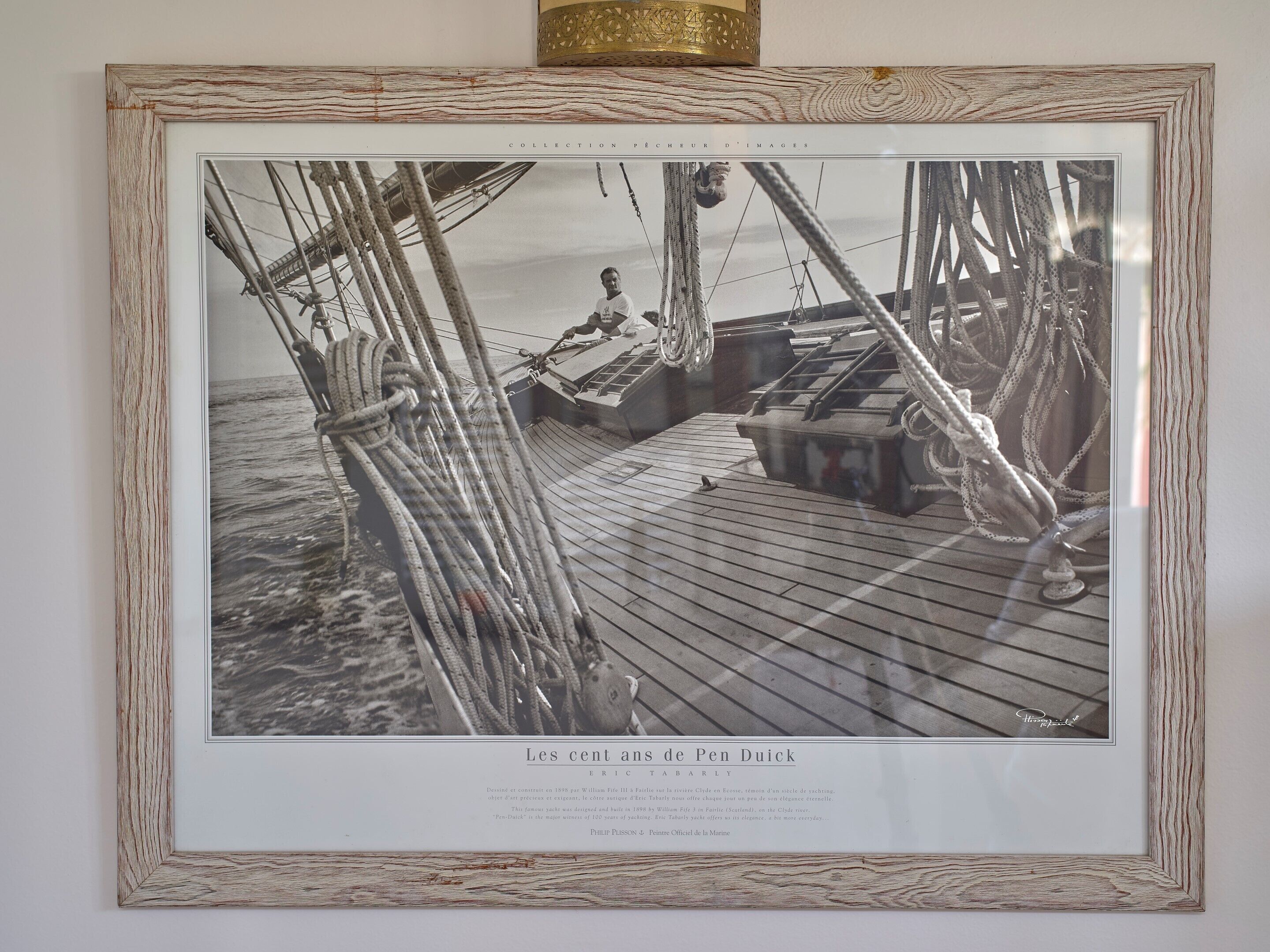 Fotografía blanco y negro enmarcada que muestra la cubierta de un barco, con cuerdas y una persona de pie.