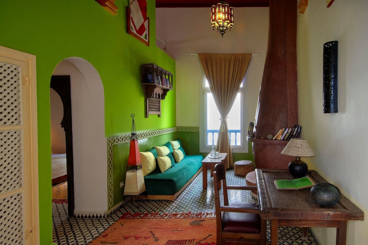 Quarto com parede verde, piso estampado, sofá verde com almofadas, mesa de madeira com luminária e um lustre.