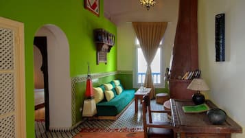 A room with a green wall, a patterned floor, a green sofa with cushions, a wooden table with a lamp, and a chandelier.