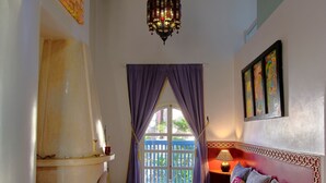 A room with a patterned floor, a bed with pillows, a hanging chandelier, and a window with purple curtains. - Ryad Watier (Essaouira)
