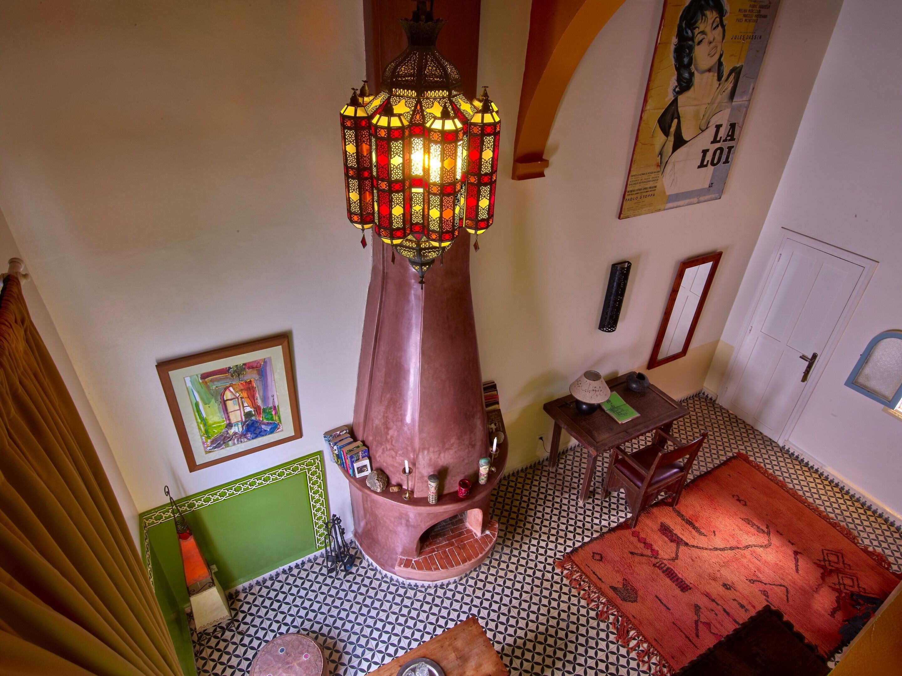 A room with a central decorative chimney, a patterned floor, a green-framed artwork, and a poster on the wall.