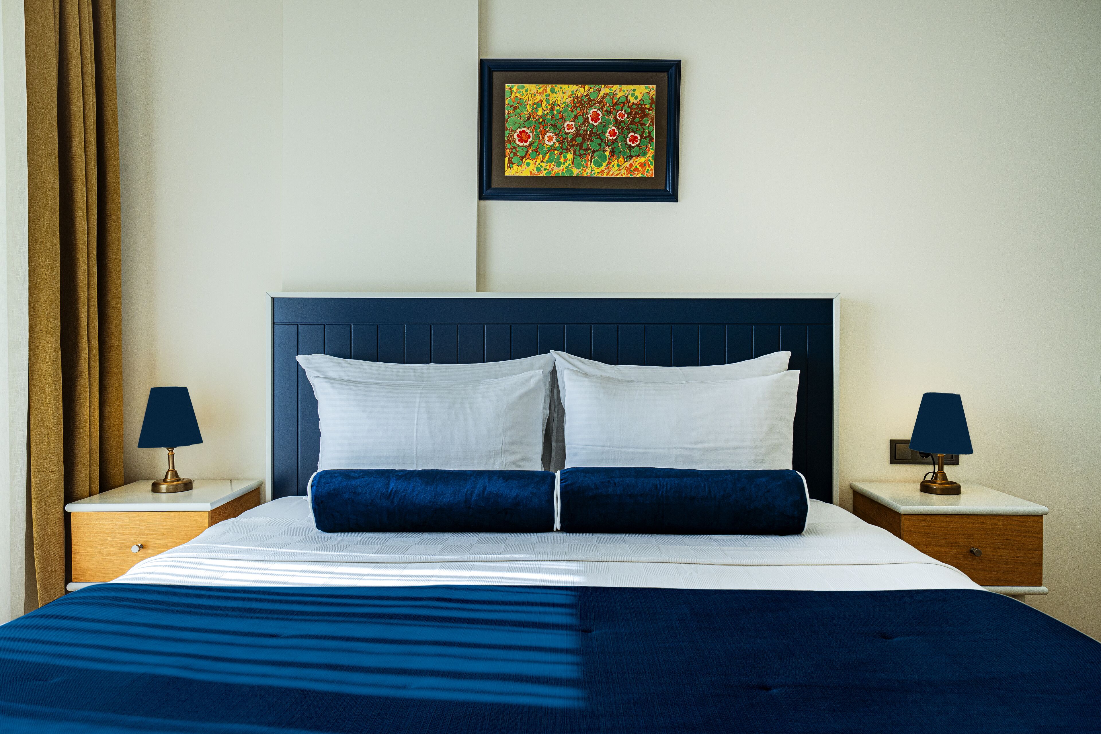 Un lit bien fait, aux couleurs bleu et blanc, deux tables de chevet avec des lampes, et une œuvre d’art encadrée accrochée au mur.