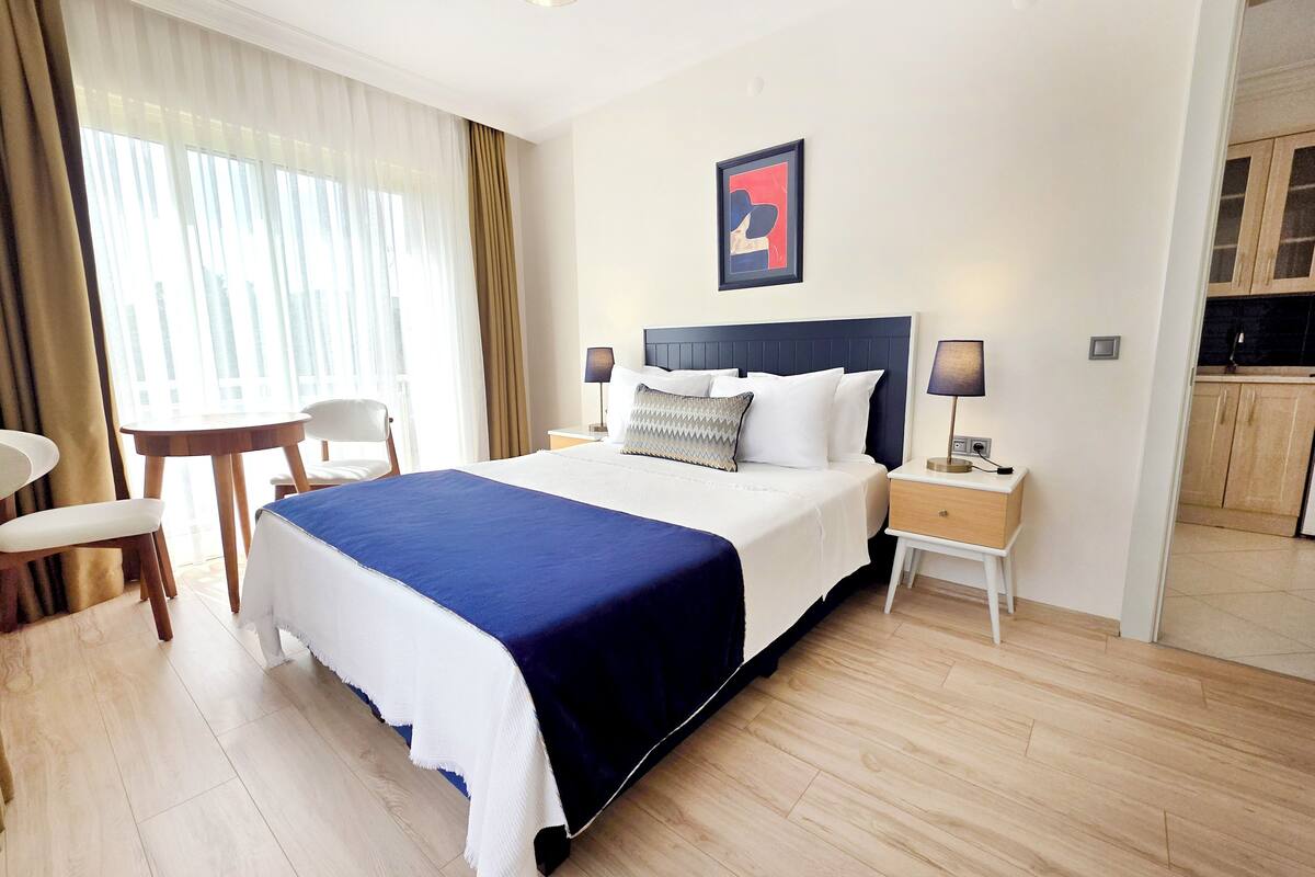 A hotel room with a bed, bedside tables, a desk, a chair, and a view of a kitchen.