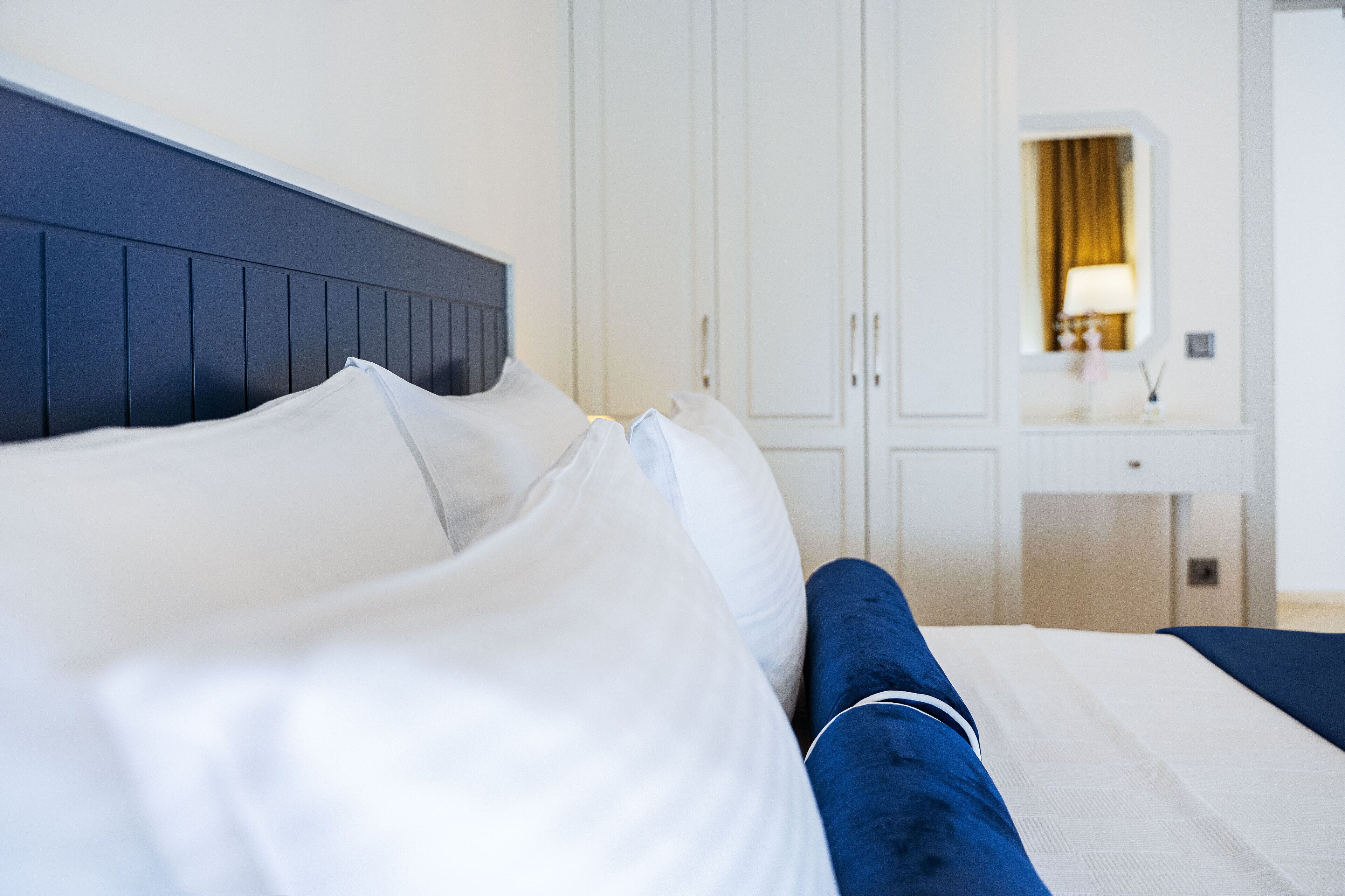 A neatly made bed with white and blue bedding, a dark headboard, and a white wardrobe with a mirror and a lamp in the background.