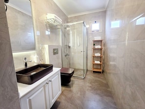 Un baño moderno con cabina de ducha de vidrio, un tocador blanco con lavabo negro y un estante de madera con toallas.