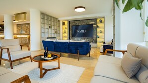 A modern living room with a blue sofa, beige armchairs, and a coffee table. A flat-screen TV is mounted on the wall, and there are shelves with decorative items. - Kamer Exclusive Hotel & Suites (Cesme)