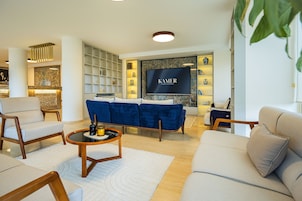 Una sala de estar moderna con un sofá azul, sillones color beige y una mesa de centro. Un televisor de pantalla plana está montado en la pared, y hay estantes con objetos decorativos.