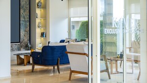 A modern hotel room with a blue sofa, a wooden coffee table, and a glass door leading to a balcony with a view of greenery. - Kamer Exclusive Hotel & Suites (Cesme)