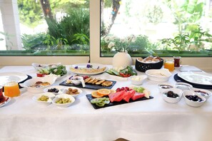 A breakfast buffet with various dishes and drinks, a vase with flowers, and a view of greenery through the window. - Kamer Exclusive Hotel & Suites (Cesme)