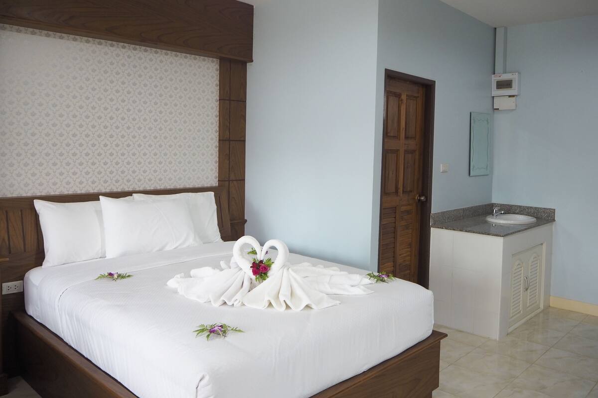 A neatly made bed with white linens and a decorative swan arrangement, a wooden headboard, a small bedside table, and a bathroom with a sink and mirror.