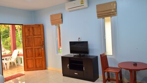 Una habitación con televisión sobre un mueble, una puerta de madera, dos ventanas con cortinas y una pequeña mesa.