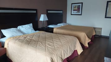 A hotel room with two beds, each with a beige quilted bedspread and white pillows. There is a nightstand with a lamp between the beds, and a framed picture on the wall.