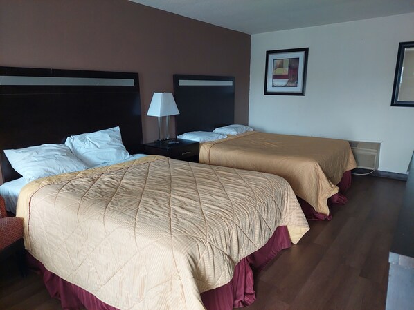 Habitación de hotel con dos camas, cada una con una colcha acolchada color beige y almohadas blancas. Hay una mesita de noche con una lámpara entre las camas, y un cuadro enmarcado en la pared.