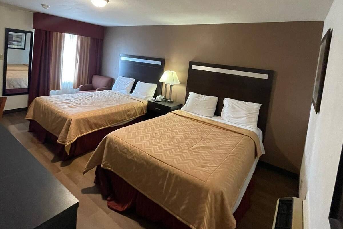 A hotel room with two beds, a desk, a chair, a nightstand with a lamp, and a framed picture on the wall.