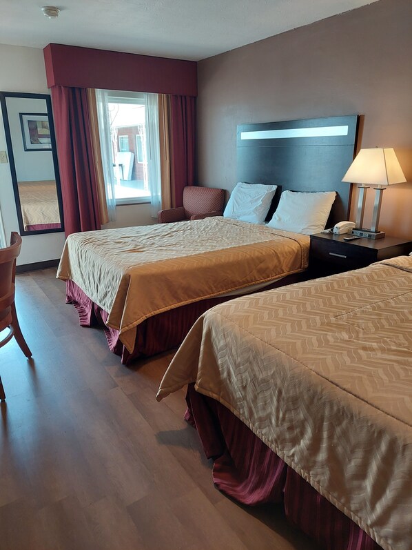 Quarto de hotel com duas camas, uma mesa de cabeceira com luminária, uma cadeira e uma janela com cortinas.