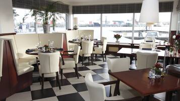 En hotelrestaurant med skakternet gulv, hvide møbler og store vinduer med udsigt over bybilledet.