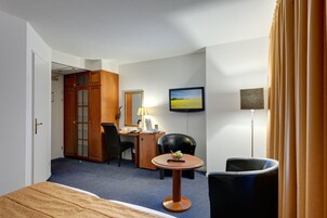 Quarto de hotel com cama, escrivaninha com luminária, cadeira, televisão e guarda-roupa.