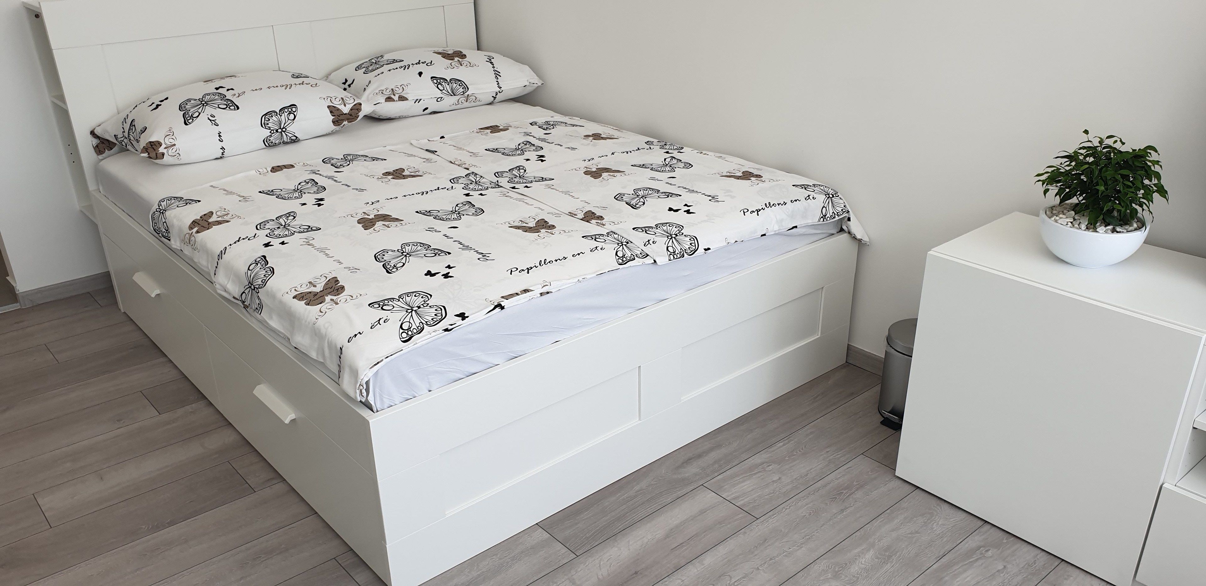 A neatly made bed with a white headboard and footboard, a white bedside table with a potted plant, and a white dresser with drawers.