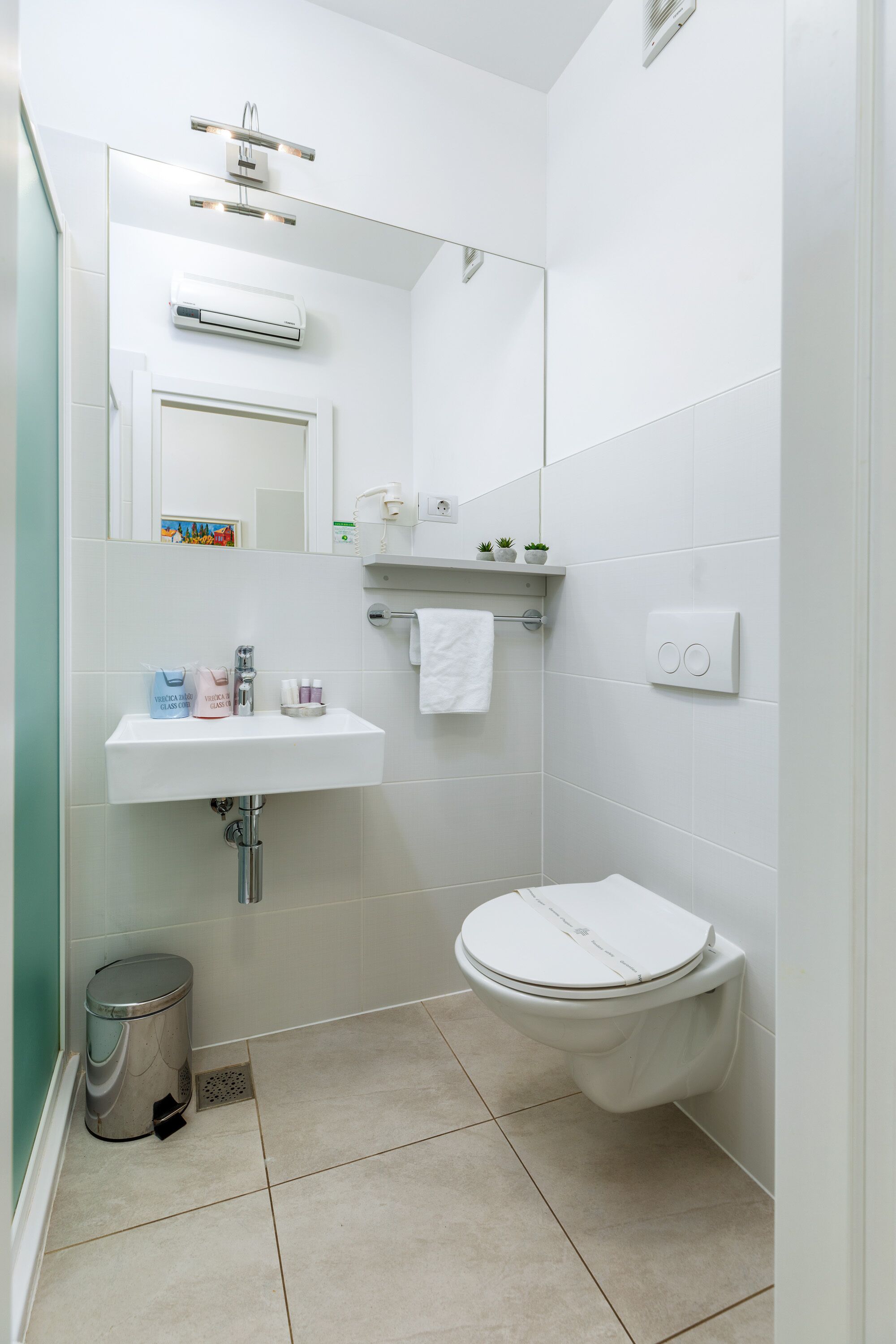 A modern bathroom with a white sink, mirror, and toilet. There is a trash can and a shelf with toiletries.