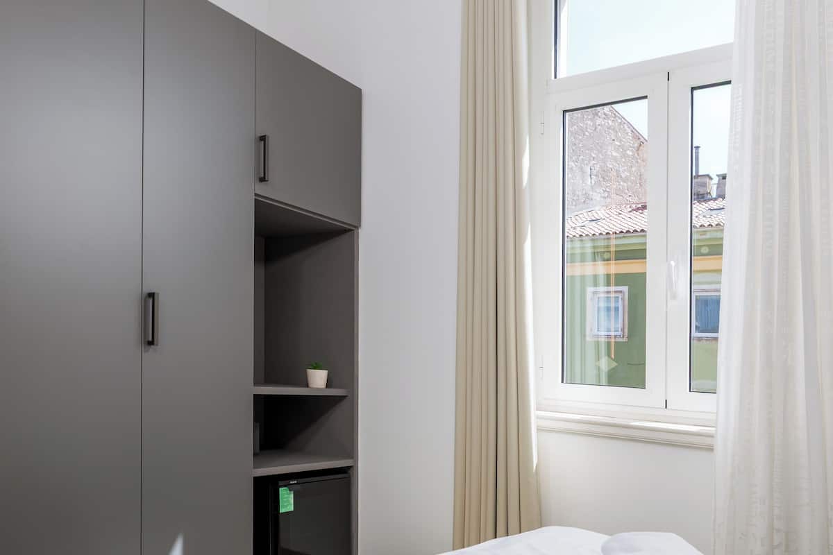 A hotel room with a bed, a wardrobe, a window with curtains, and a view of buildings outside.