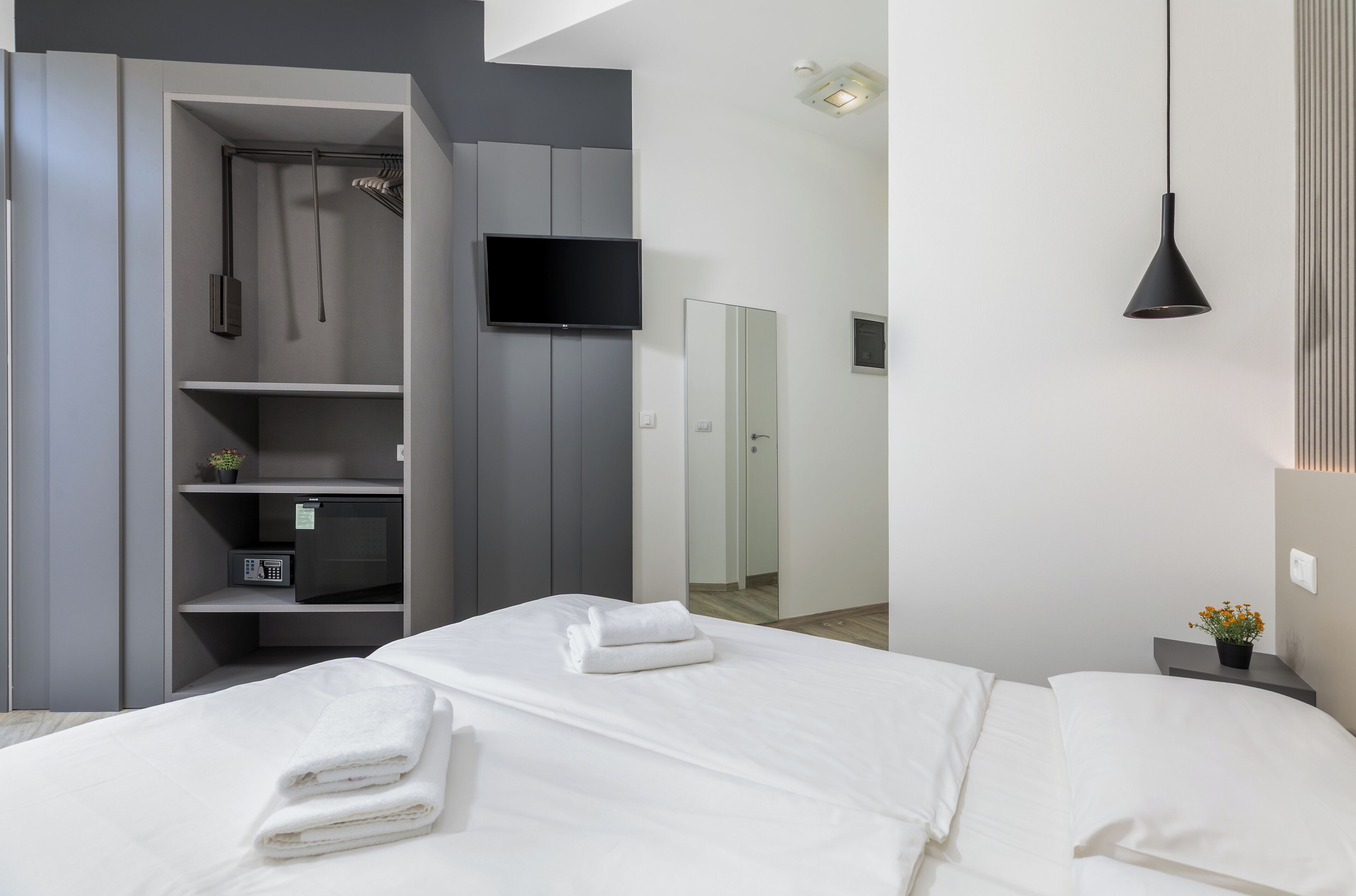 Una habitació d'hotel moderna amb un llit, un armari, una televisió i una taula de nit amb una planta.