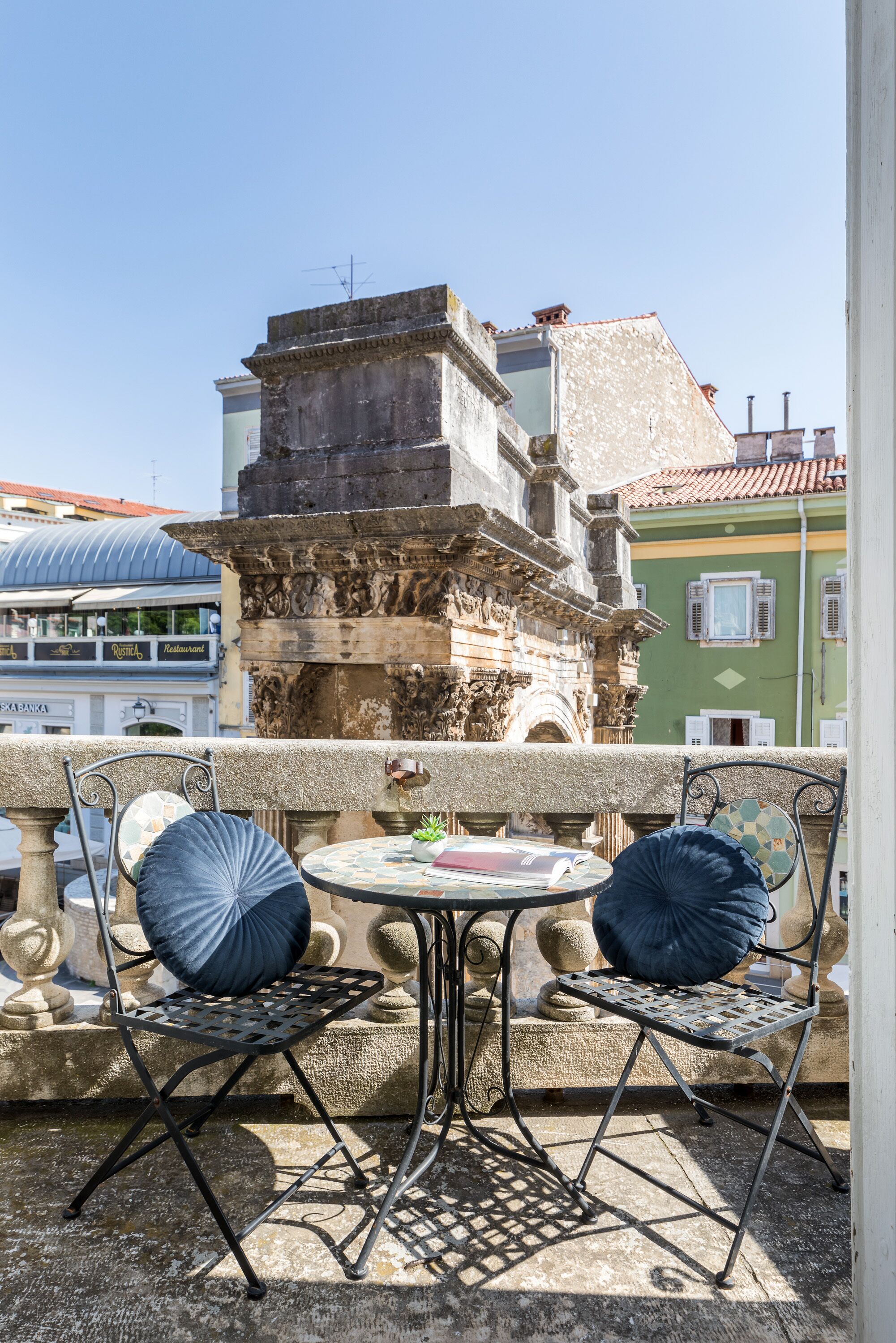 Ein Balkon mit zwei Metallstühlen und einem kleinen runden Tisch, mit Blick auf eine historische Steinarchitektur mit aufwendigen Schnitzereien.