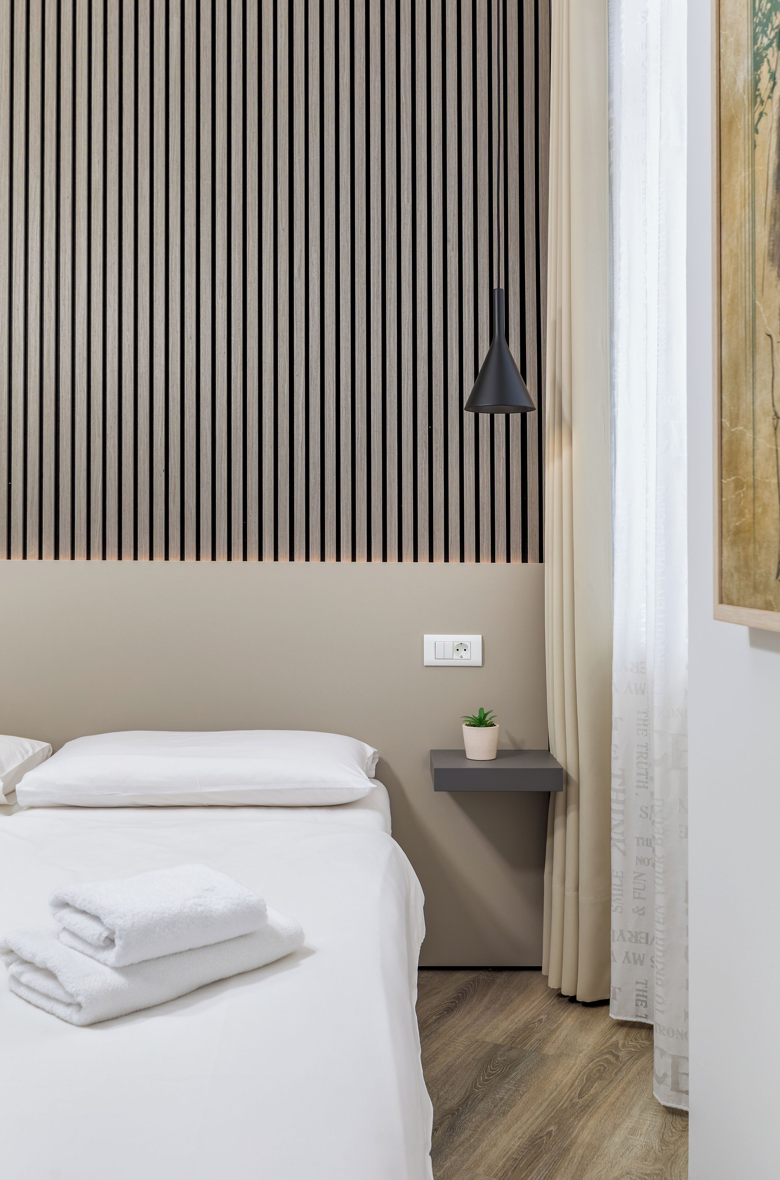 A neatly made bed with white linens, a small bedside table with a plant, a wall-mounted light, and a power outlet.