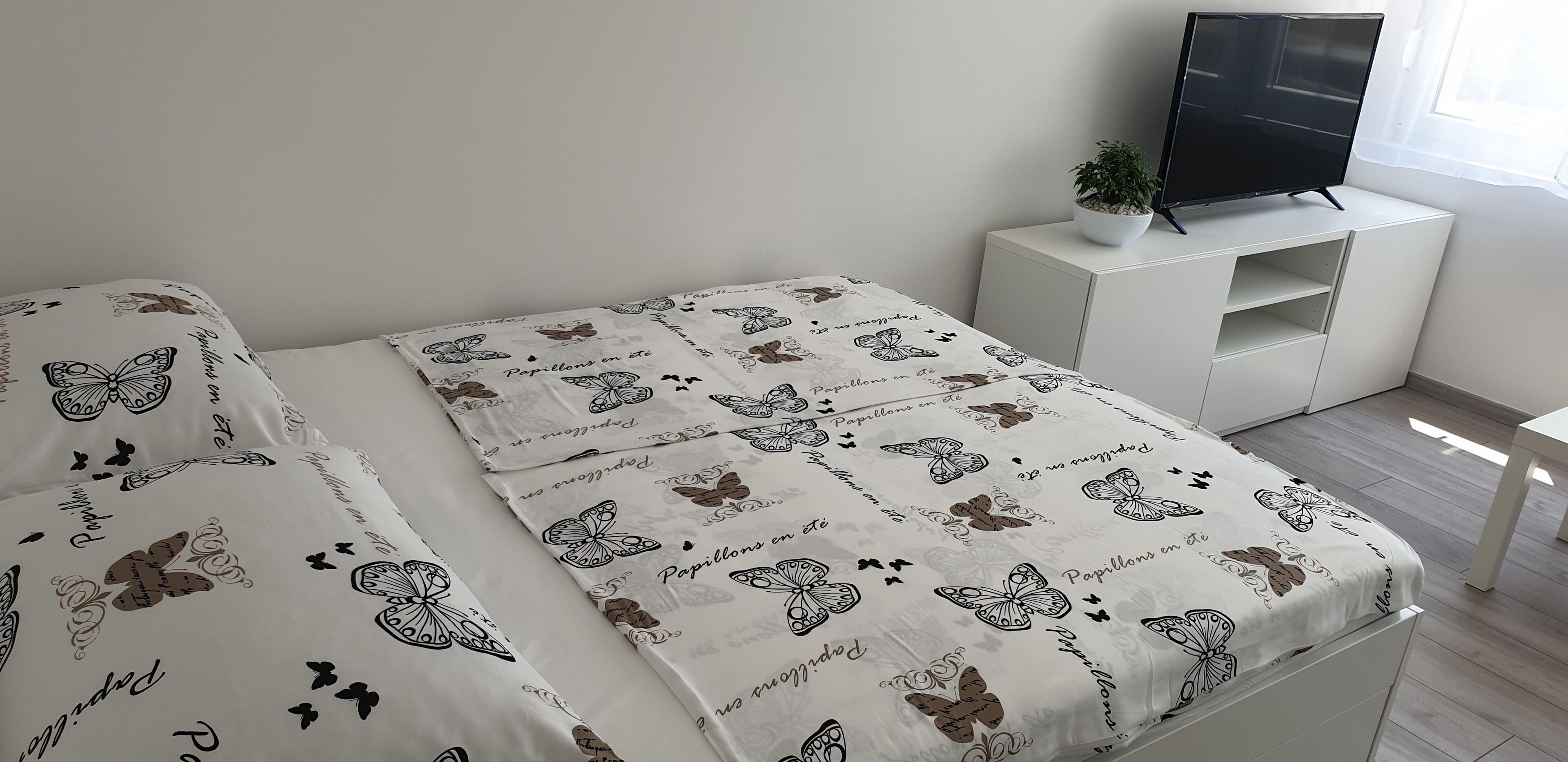 Uma cama bem arrumada com colcha decorada com tema de borboletas, uma televisão sobre um suporte branco e um vaso de planta.