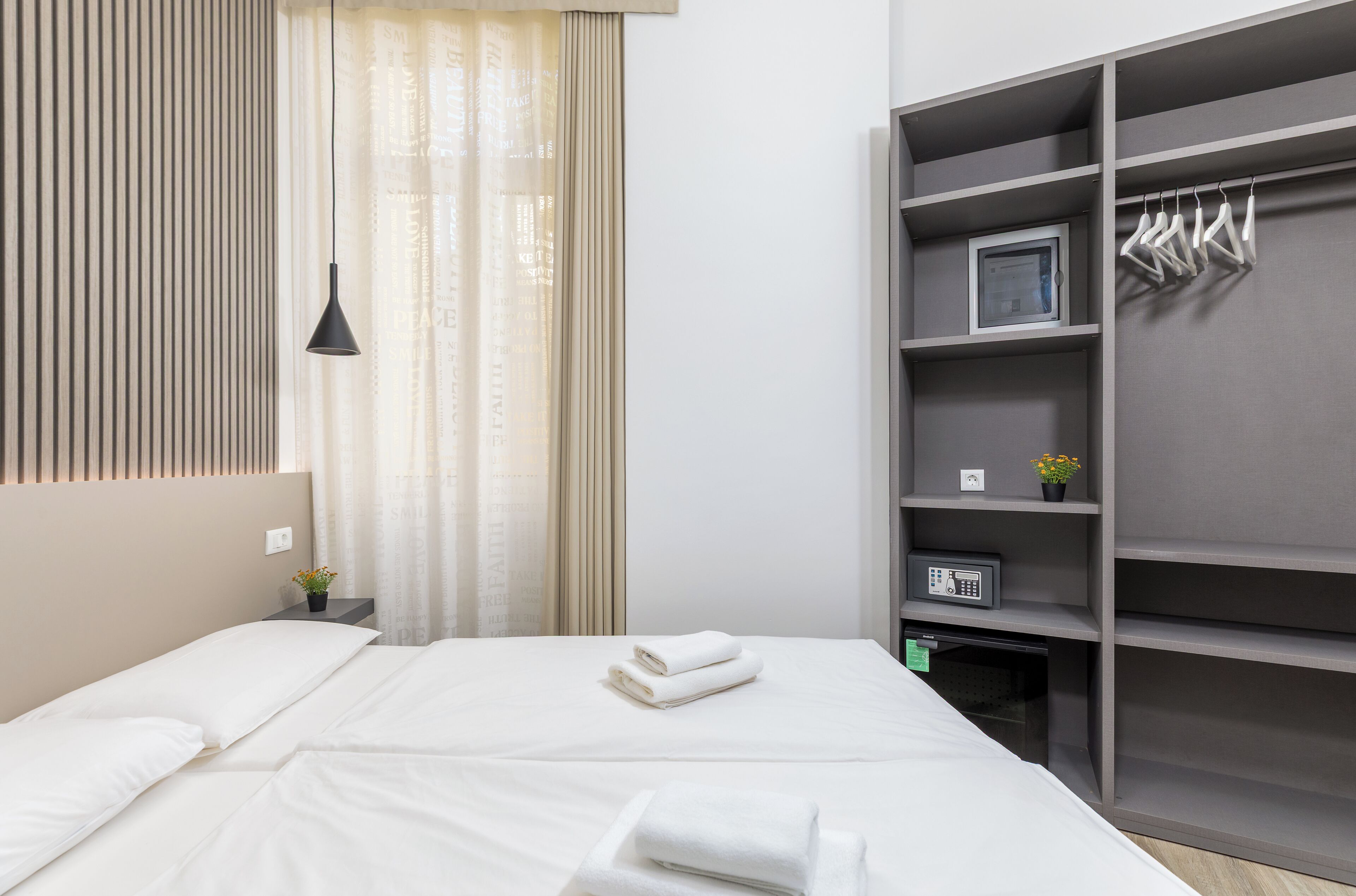 A neatly made bed with white linens, a bedside table with a lamp, and a gray wardrobe with hangers.