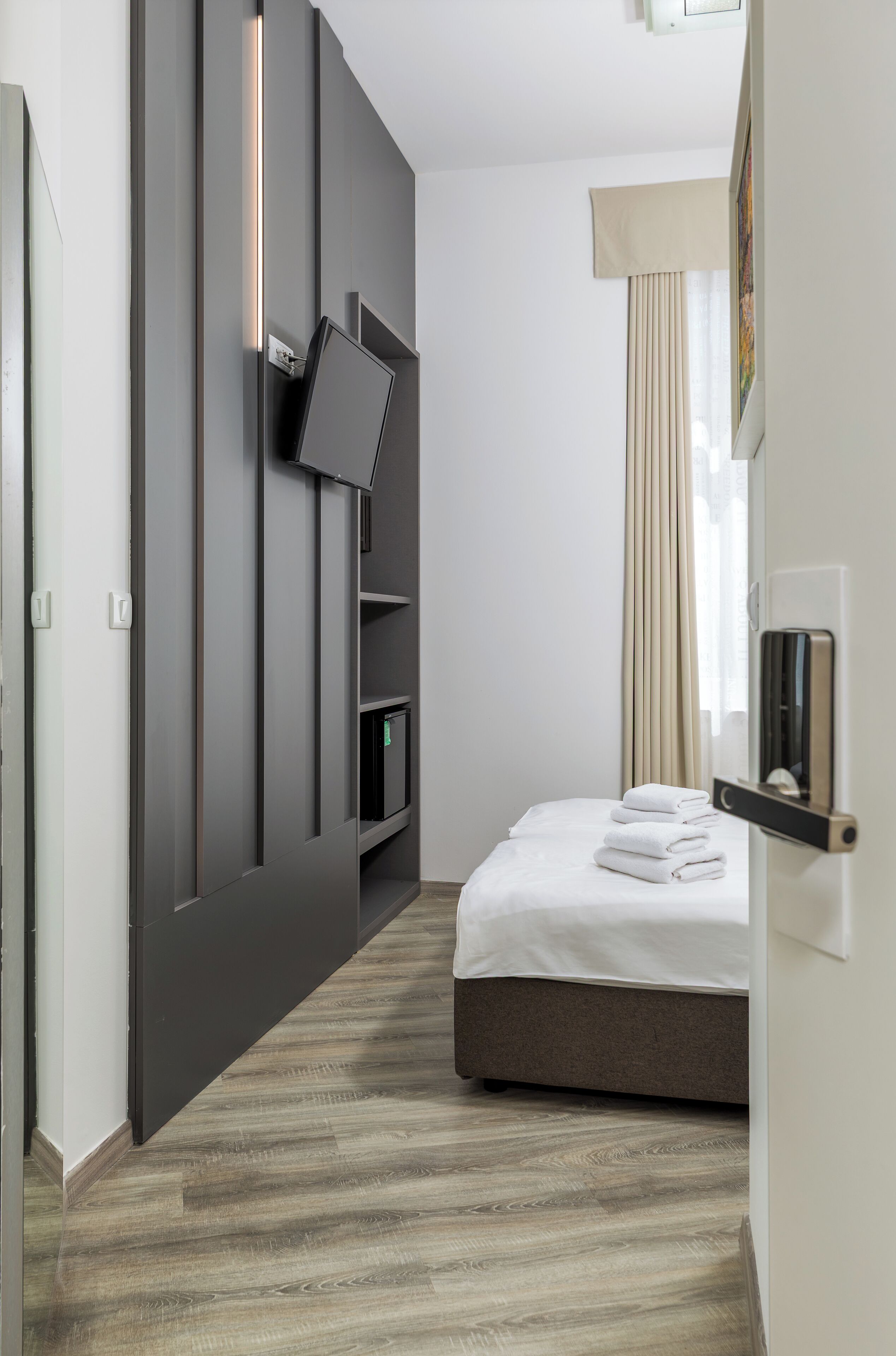 A modern hotel room with a flat-screen TV mounted on a dark wall, a bedside table with a lamp, a bed with a beige headboard, and a window with curtains.