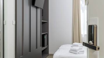 Habitación de hotel moderna con televisor de pantalla plana montado en una pared oscura, una mesita de noche con lámpara, cama con cabecera color crema y una ventana con cortinas.