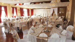 Un gran saló de banquetes amb taules rodones preparades per a un esdeveniment formal, amb teles de taula blanques, detalls daurats i una presentació de taula elegant.