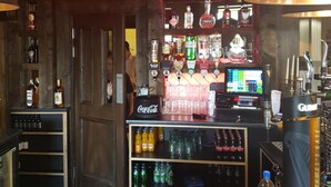 Zona de bar amb un cartell de neó que diu "THE COHANNONINN". Hi ha prestatges amb diverses ampolles i vasos, un nevera de Coca-Cola i una pantalla digital que mostra informació.