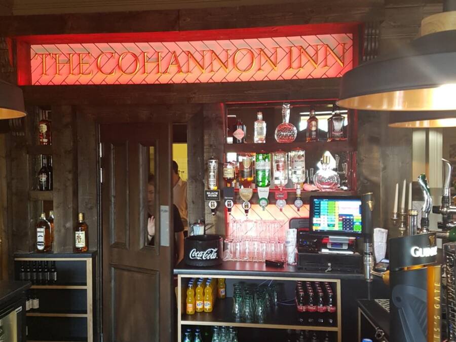 Un espace bar agrémenté d’un panneau néon sur lequel on peut lire « THE COHANNONINN ». On y trouve des étagères remplies de bouteilles et de verres de toutes sortes, un réfrigérateur à boissons Coca-Cola et un écran numérique affichant des informations.