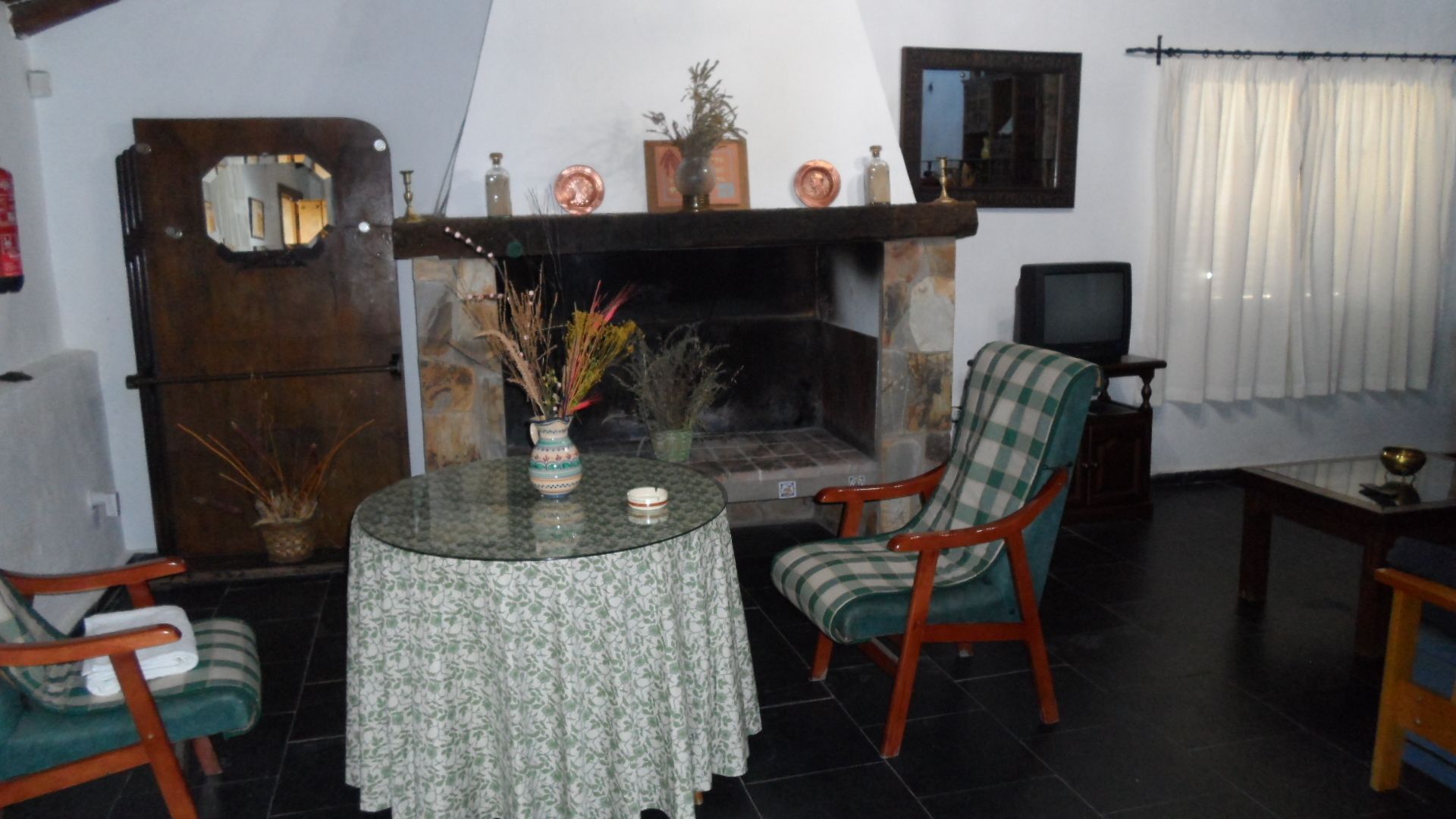 Una sala de estar con chimenea, una mesa redonda con mantel estampado, dos sillones y un televisor sobre un mueble.