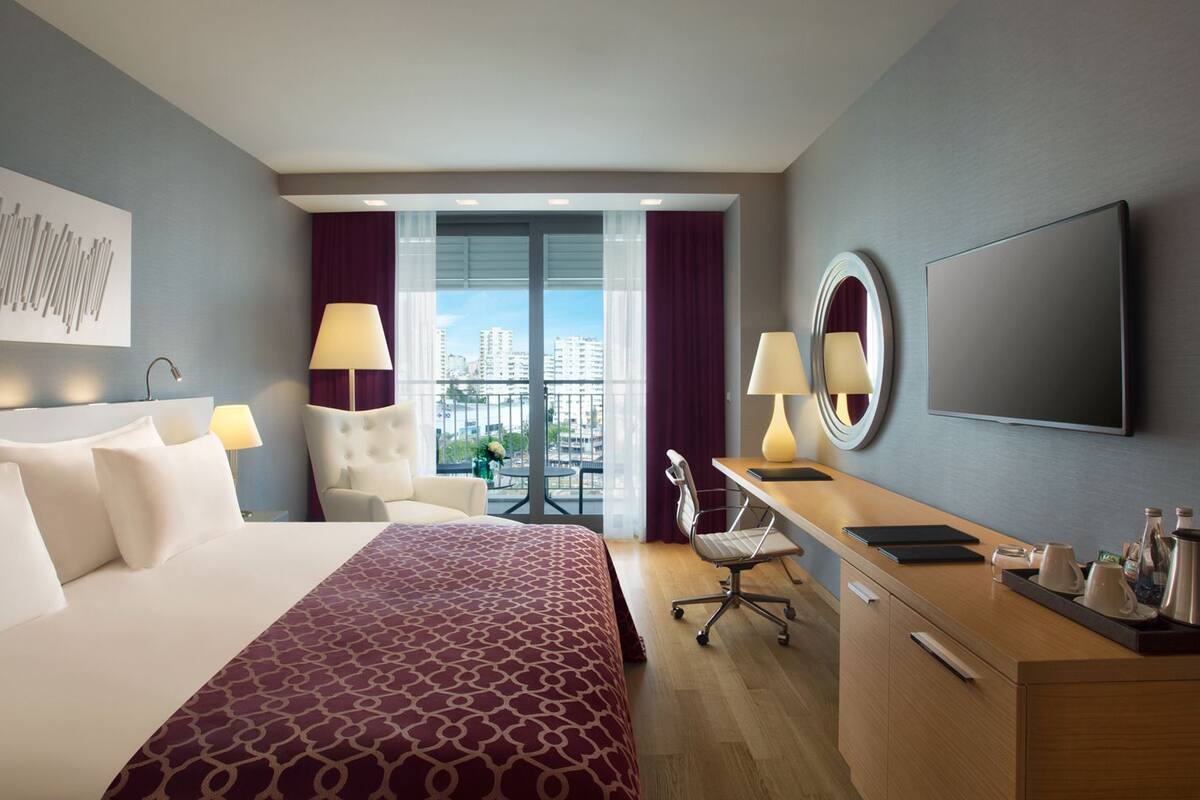 A hotel room with a large bed, a desk with a chair, a TV, and a view of the city through the window.