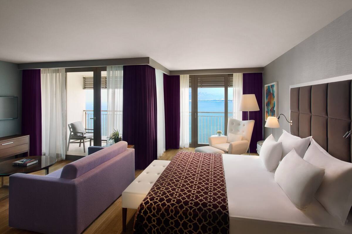 A modern hotel room with a large bed, a sofa, a TV, and a balcony with curtains.
