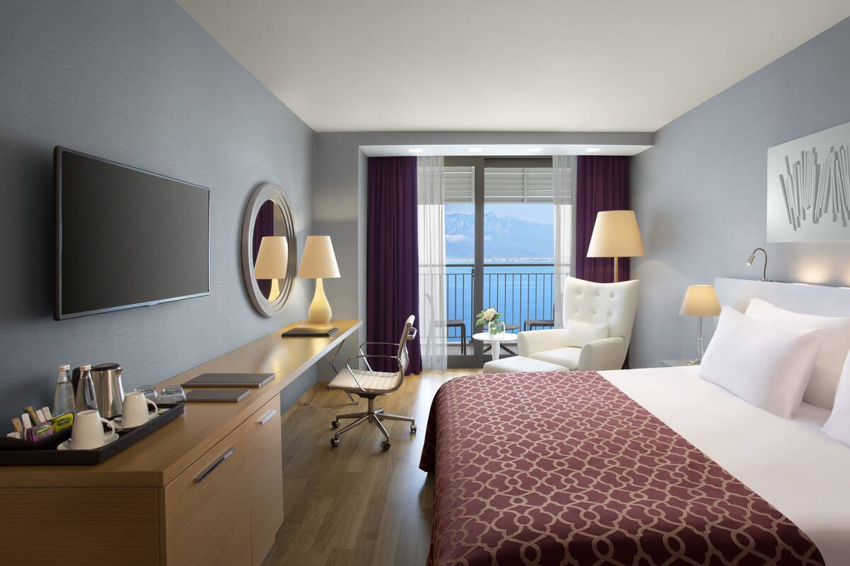A hotel room with a bed, a desk with a laptop, a chair, a TV, and a view of the sea.