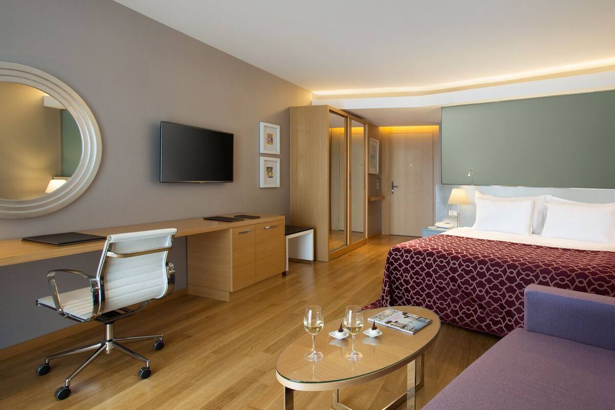 A hotel room with a bed, a desk, a chair, a TV, and a round table with glasses and a magazine.