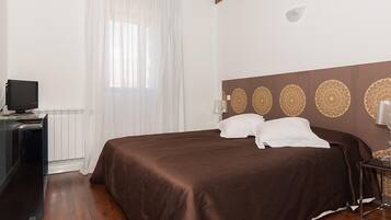 A bedroom with a large bed, a television on a desk, and a window with sheer curtains.