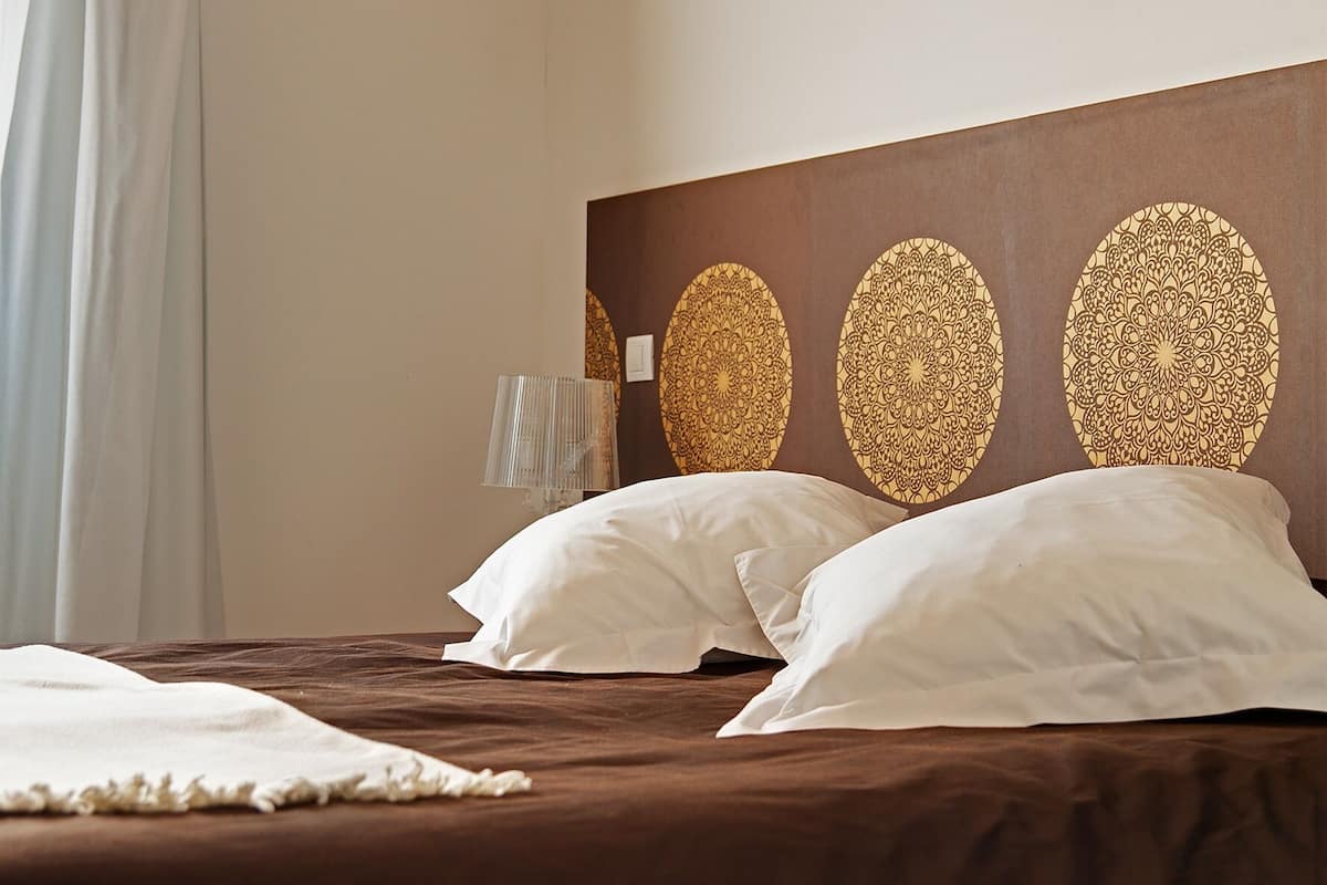 Habitación de hotel con una cama grande, dos almohadas blancas, cabecera marrón con paneles decorativos y una lámpara de noche.