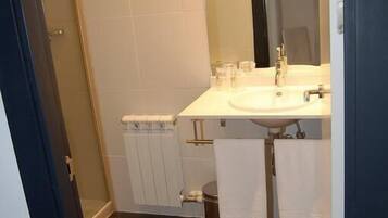 Baño con lavamanos blanco, espejo y toallero. Se ve un inodoro y una zona de ducha.