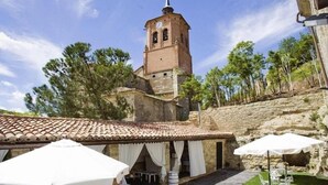 Un patio con paredes de piedra, techo de tejas, mobiliario exterior con sombrillas y una torre del reloj al fondo.