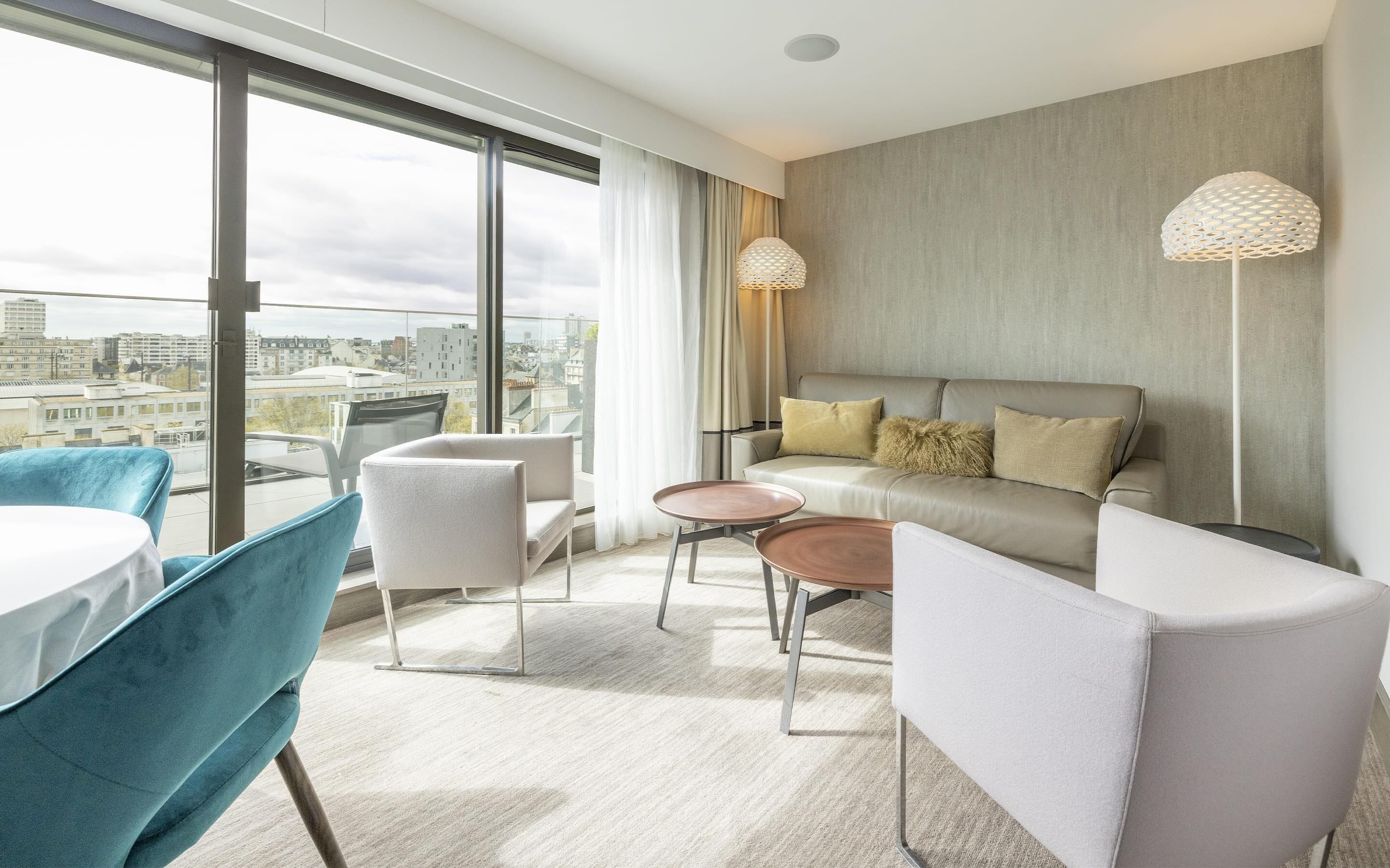 A modern hotel room with a large window, a sofa, a dining table, and two chairs.