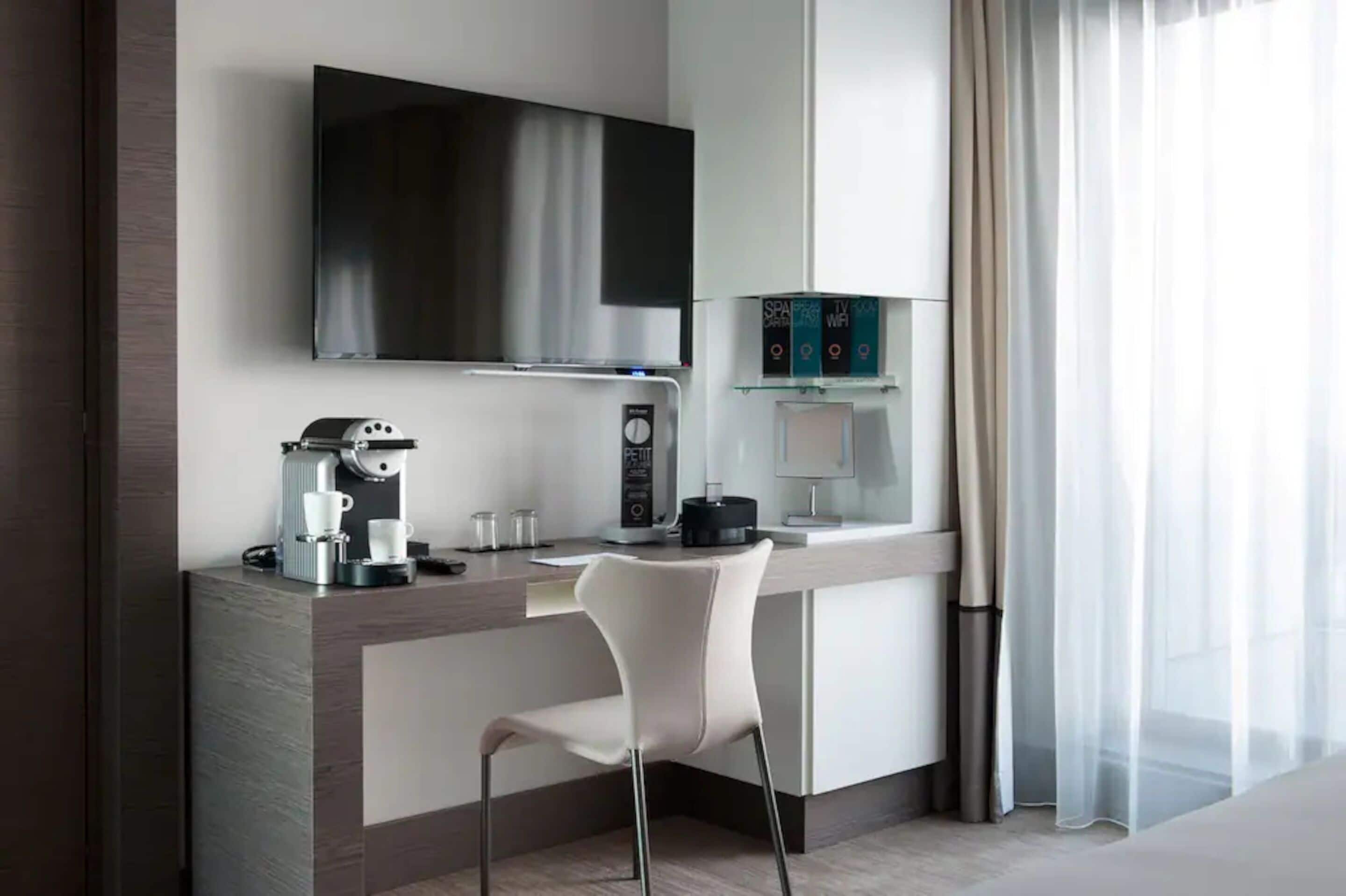 Habitación de hotel moderna con una televisión de pantalla plana sobre un escritorio de madera, una cafetera, una silla blanca y una ventana con cortinas translúcidas.