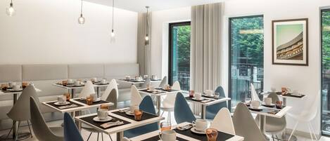 A modern dining area with multiple tables and chairs, large windows with a view of greenery, and a ceiling with recessed lighting.