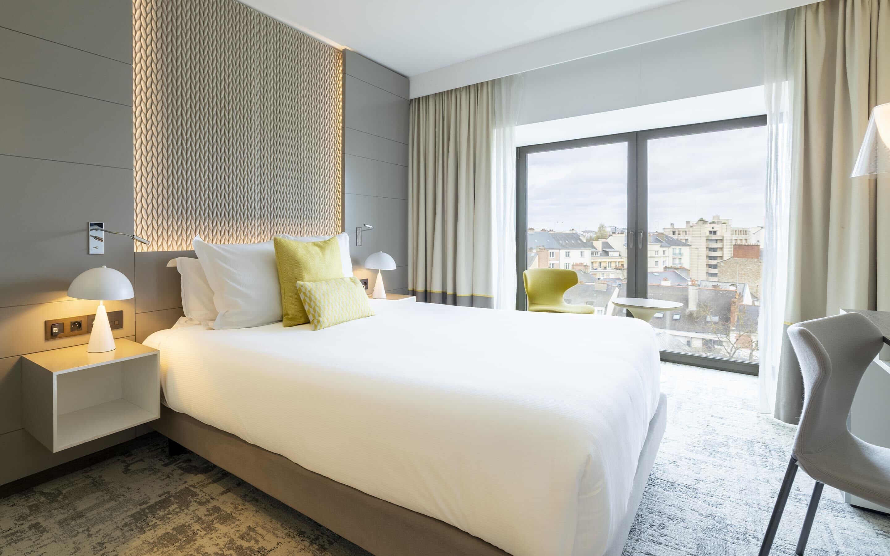 A hotel room with a large bed, a nightstand with a lamp, a chair, and a view of the city through a large window.