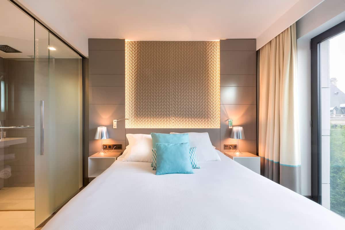 A modern hotel room with a large bed, two bedside tables, a wall-mounted light panel, and a view of the outdoors.