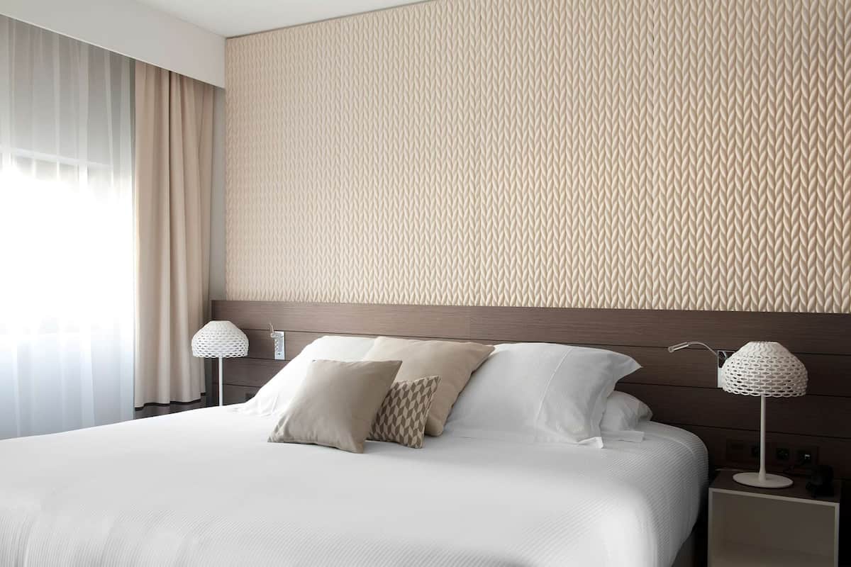 A neatly made bed with white linens and two pillows, a bedside lamp, and a wall with a geometric pattern.