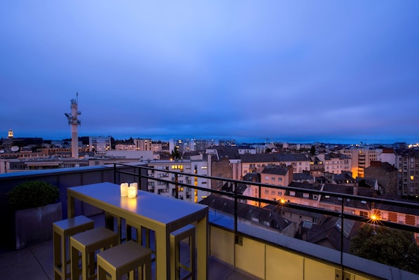 Terraza en la azotea con una mesa y sillas, con vista a un paisaje urbano al atardecer.